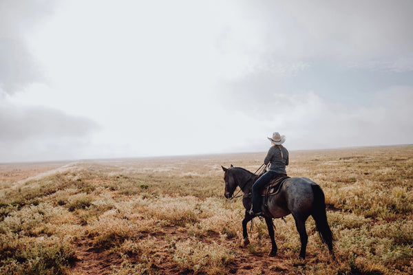 Cowgirl riding a horse in a vast, open field on a cloudy day