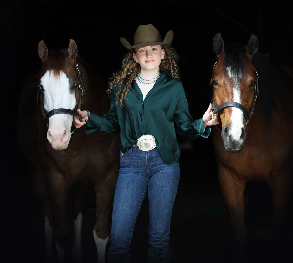 Woman in a green shirt and cowboy hat standing between two brown horses against a dark background